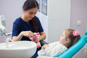 Pediatric dentist educating a smiling little girl about proper tooth-brushing, demonstrating on a model. Early prevention, raising awareness, oral hygiene demonstration concept