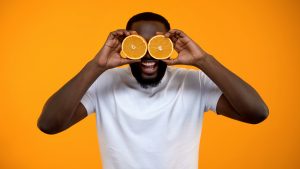 Joyful Afro-American man closing eyes with half of oranges, vitamins for health